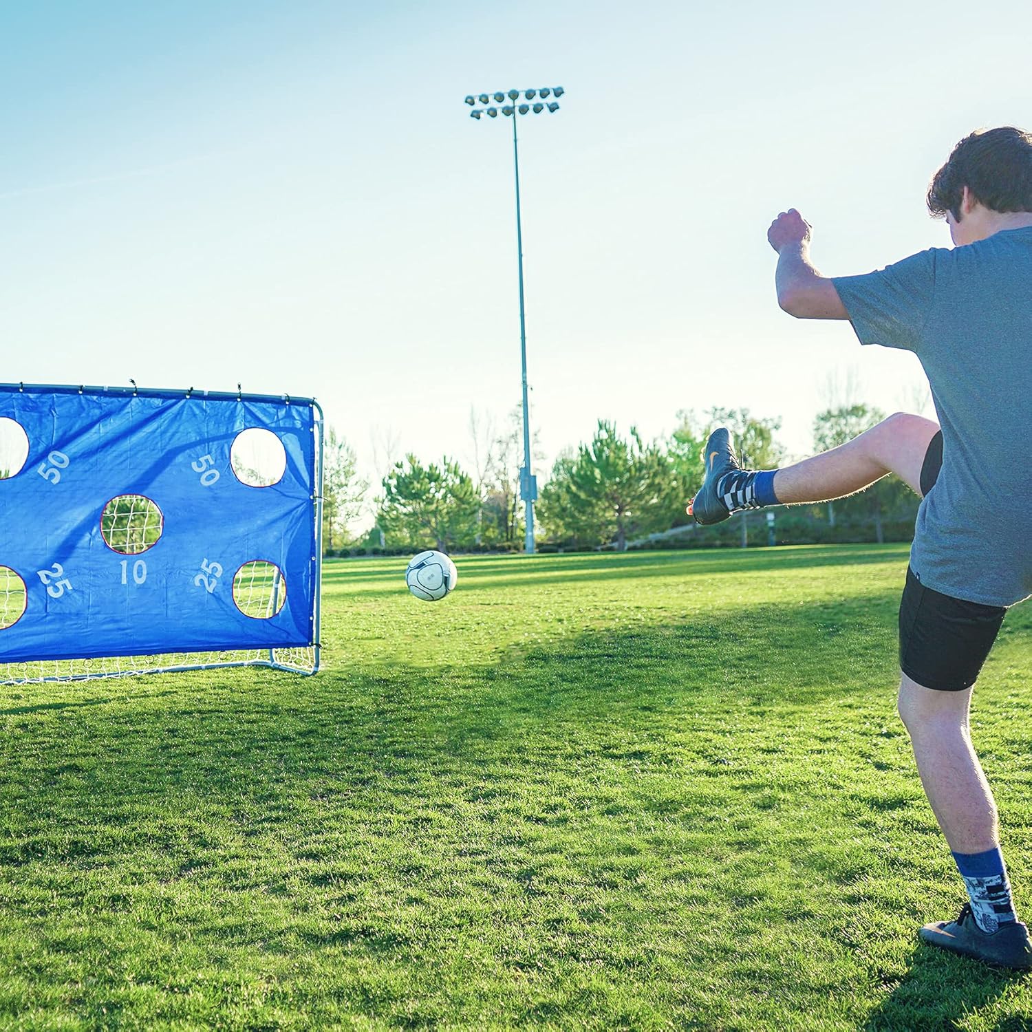 Person kicking a soccer ball towards a target on a grassy field with trees in the background.